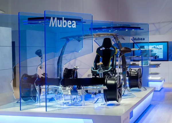 Futuristic car chassis display with transparent panels, showcasing wheels, seat, and steering components at a Mubea exhibition booth.