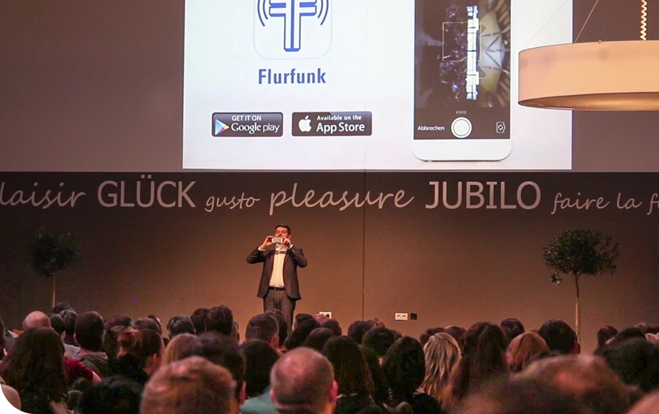 A speaker presents on stage to a large audience, with a screen displaying a smartphone app and download options from Google Play and the App Store.