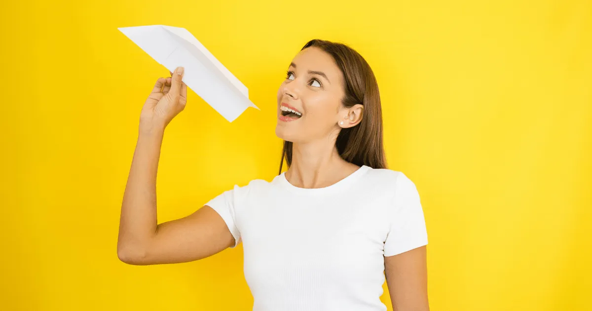 Smiling woman in a white t-shirt holding a paper airplane, preparing to throw it, against a bright yellow background.