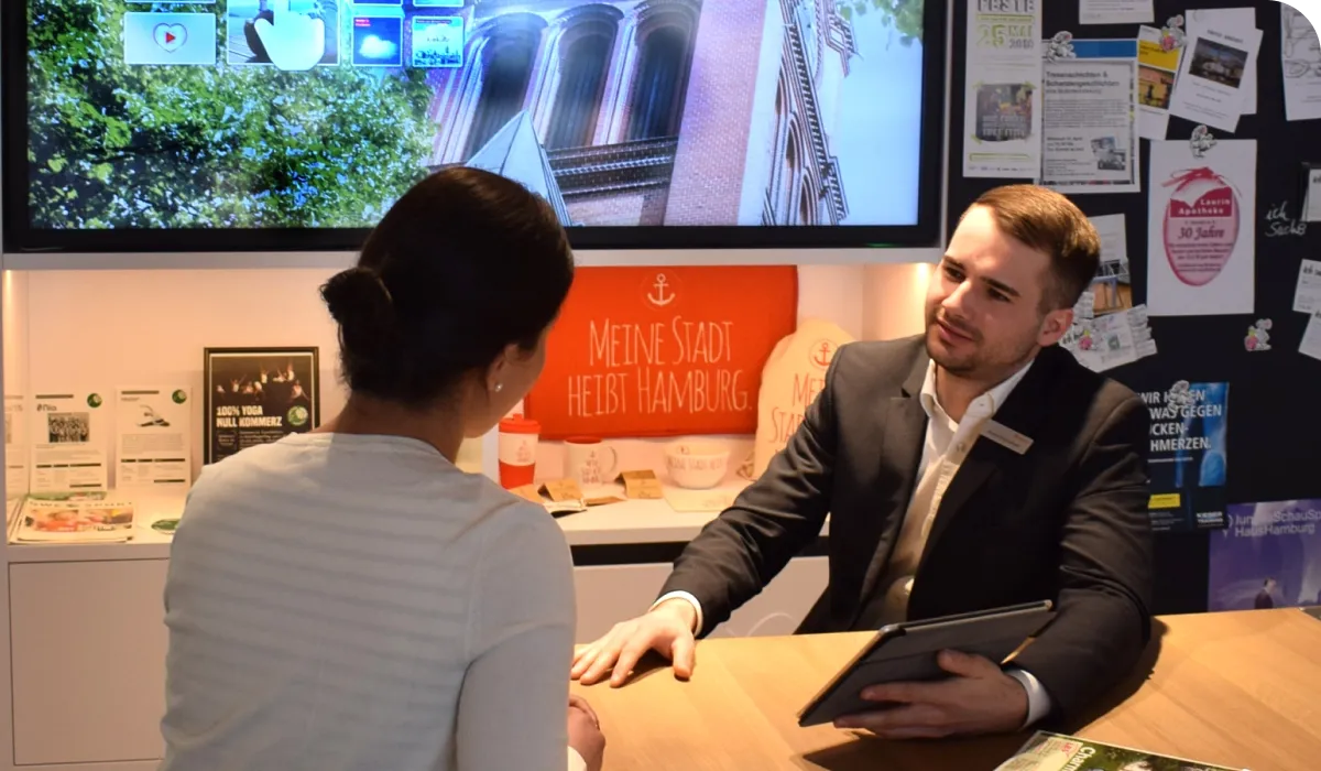 A man in a suit holds a tablet while talking to a woman at a desk. A large screen and various posters are visible in the background.