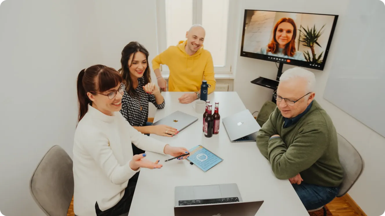 Four Staffbase colleagues in a meeting room with laptops, joined by a remote colleague on a video screen.