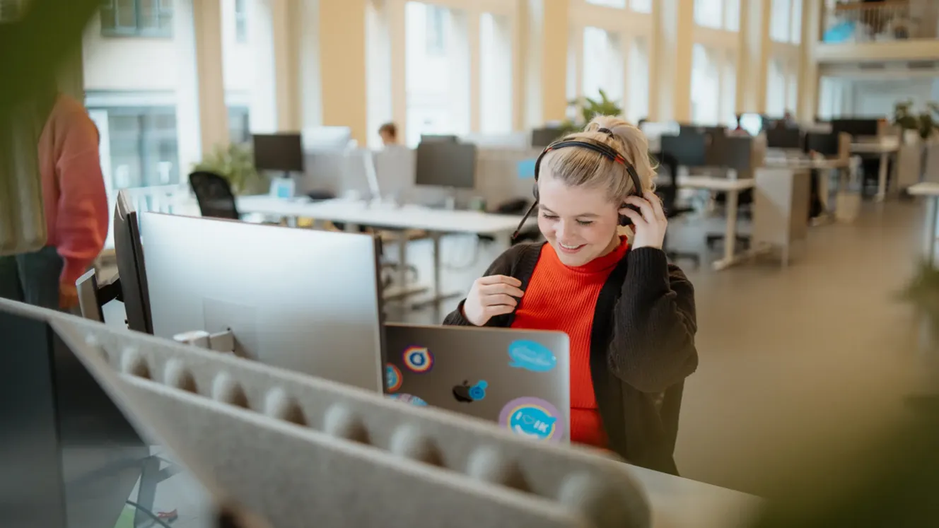 Staffbase employee smiling while adjusting her headset at her desk in an open-plan office.