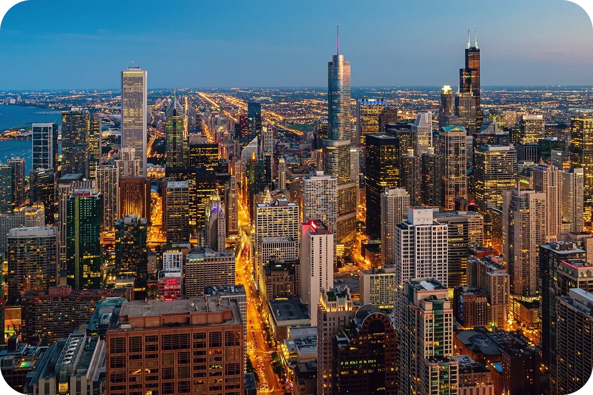 Aerial view of a cityscape at dusk, featuring illuminated skyscrapers, streets, and a vast expanse of buildings extending to the horizon.