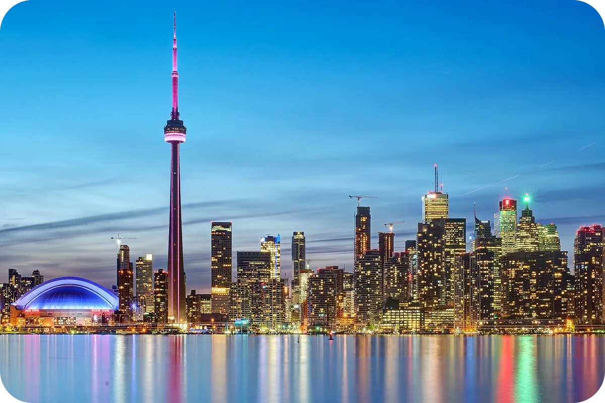 Toronto skyline at dusk with the CN Tower illuminated, surrounded by skyscrapers and their reflections shimmering in the calm water.