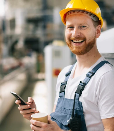 Smiling construction worker in a yellow hard hat, holding a smartphone and a cup, stands in an industrial setting.