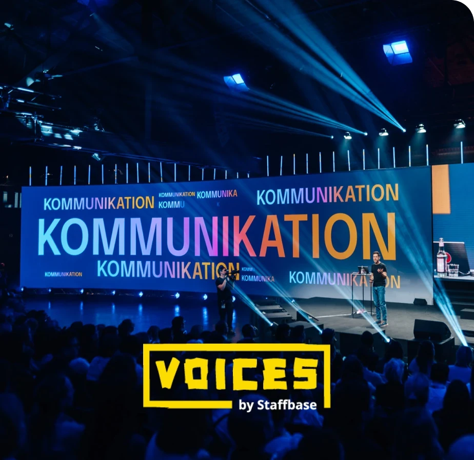 A speaker on stage at a conference with "Kommunikation" displayed in bold letters on a screen. The event is titled "Voices by Staffbase."