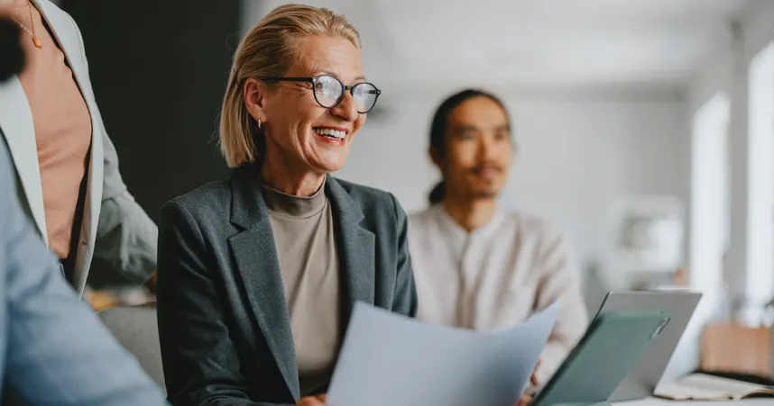 woman in glasses and blazer holding paper and smiling with coworkers surrounding her