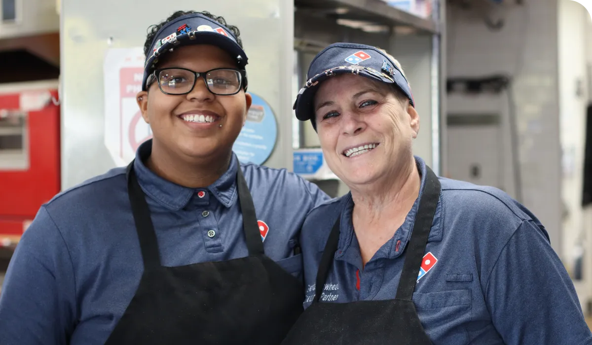 Two smiling Domino's employees in uniform, including hats and aprons, standing together in a kitchen setting.