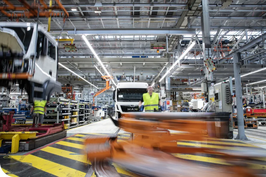 A busy factory floor with workers assembling trucks. A worker in a yellow vest stands near a truck cab, with machinery and equipment around.