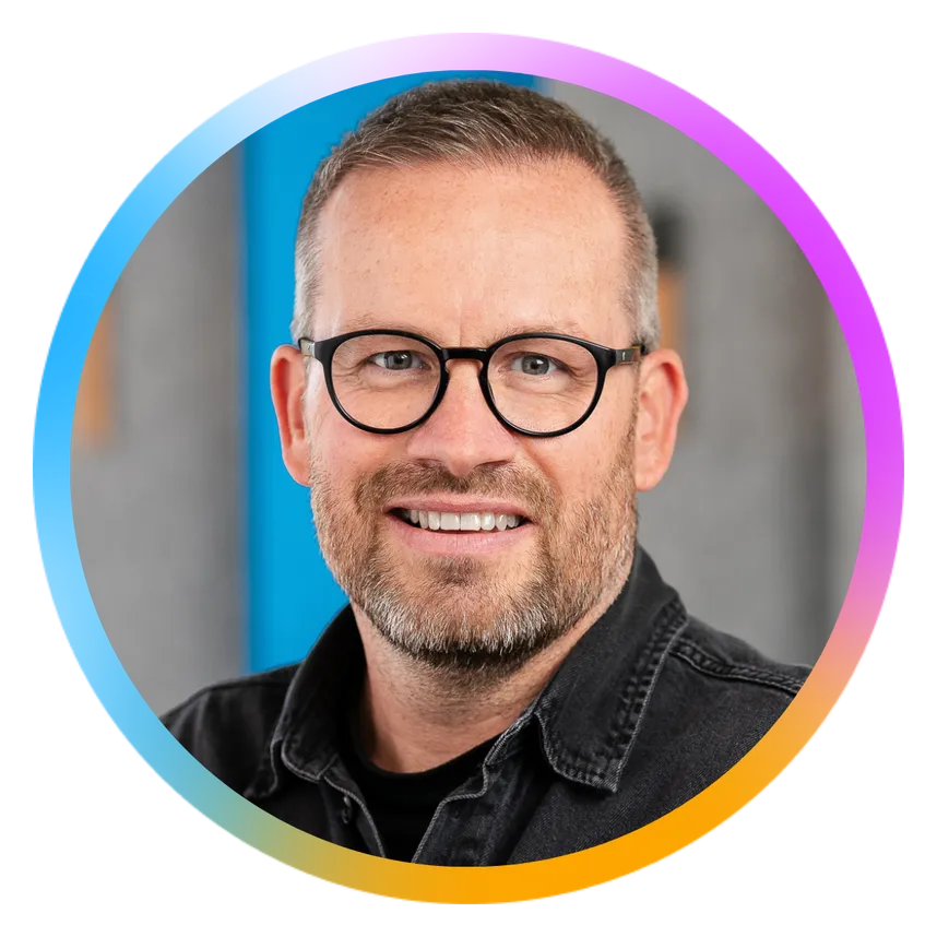 Man with glasses and a beard smiling, wearing a black shirt. Colorful circular border around the image.
