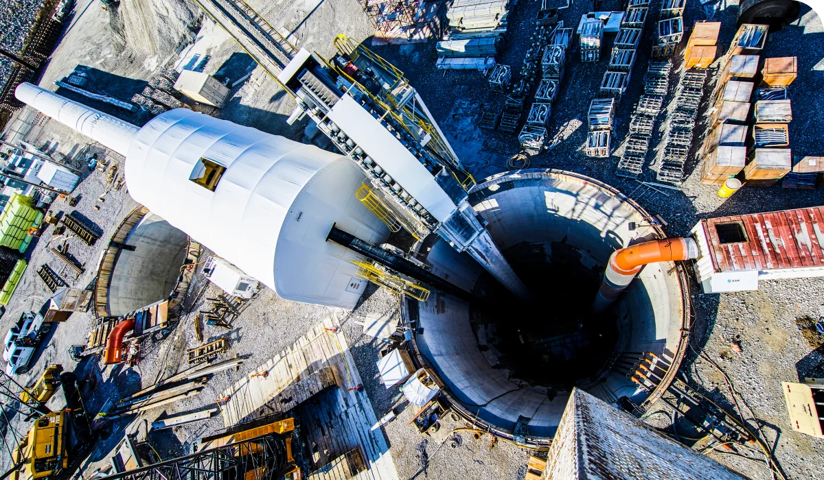 SAK. Aerial view of a large construction site with a tunnel boring machine positioned over a deep circular shaft, surrounded by equipment and materials.