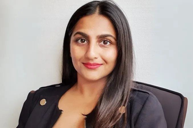 A woman with long dark hair and a black jacket smiles softly, sitting against a plain white background.