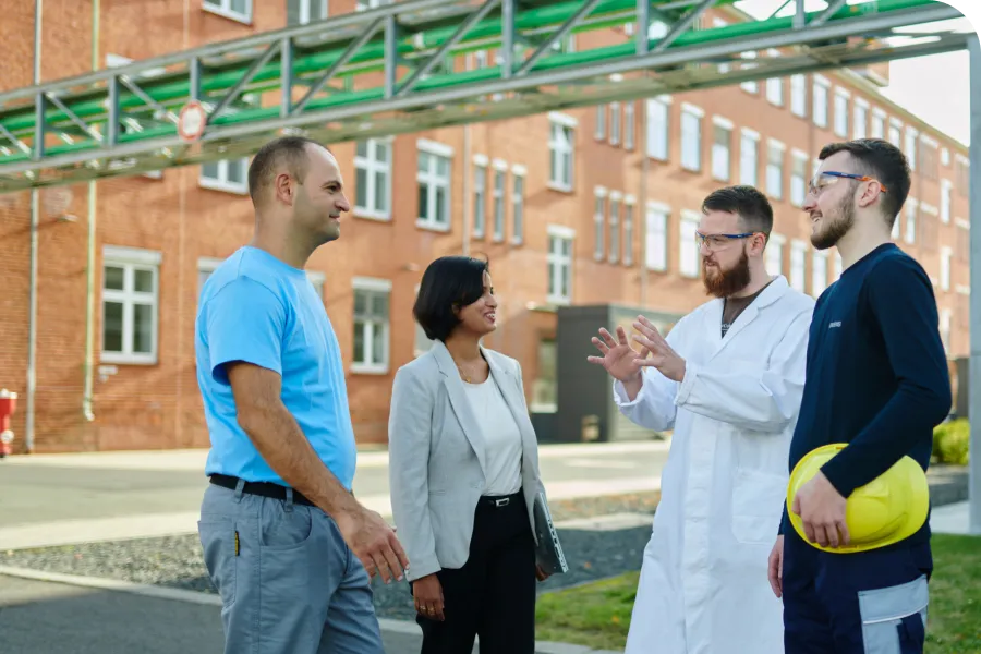 Four professionals, including a woman and three men, converse outdoors near a brick building. One wears a lab coat, another holds a yellow hard hat.