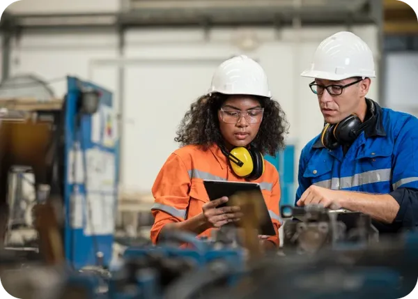 Two factory workers in hard hats and safety gear review a tablet next to industrial machinery.