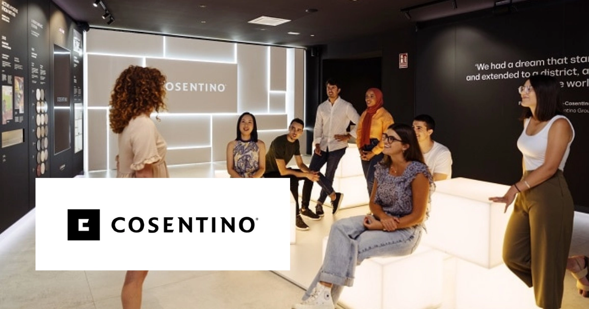 A group of people sitting and standing in a modern showroom with illuminated displays, listening to a presenter. Cosentino logo is visible.