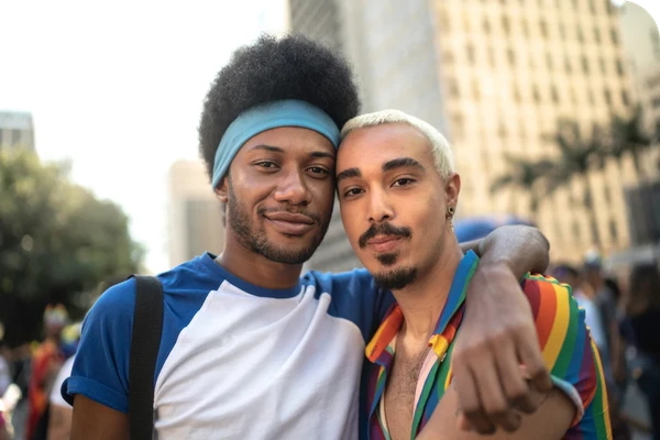 Two people smiling and embracing outdoors, one wearing a blue headband and shirt, the other in a rainbow-striped shirt, with buildings in the background.