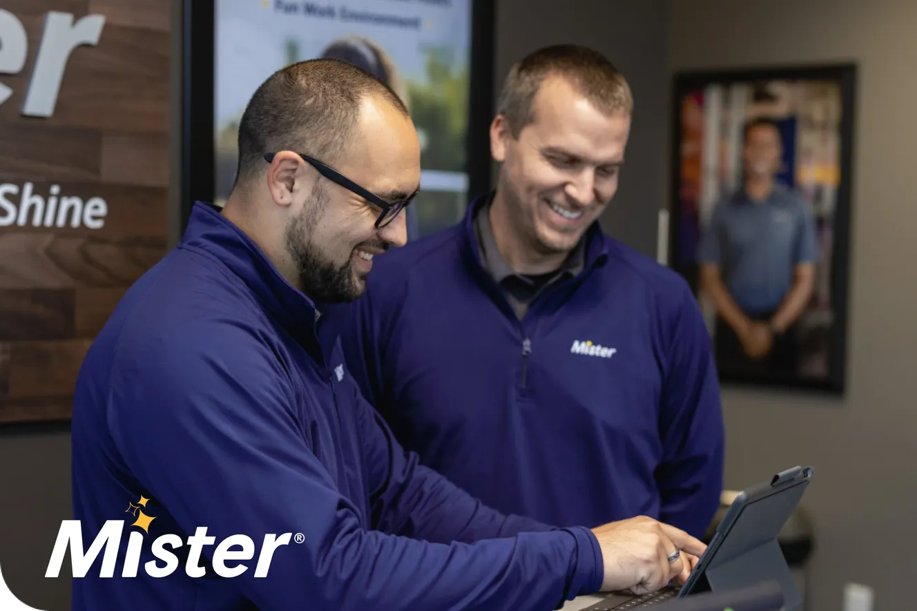 Two Mister Car Wash employees in navy branded jackets smiling and reviewing content together on a tablet at a checkout counter