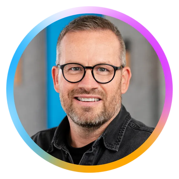 Man with glasses and a beard smiling, wearing a black shirt. Colorful circular border around the image.