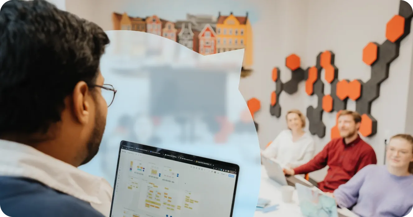 Staffbase employee reviewing a Miro board on a laptop, with colleagues discussing in a meeting room in the background.