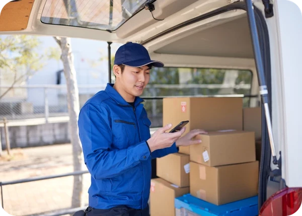 Delivery worker in blue uniform checks his phone at an open van loaded with stacked cardboard boxes.