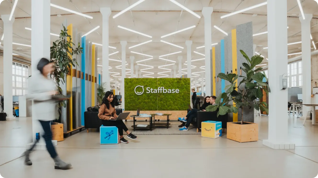 Staffbase office lounge with a green moss logo wall, employees working on laptops and chatting on black sofas.