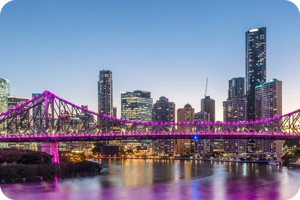 City skyline at dusk with a brightly lit bridge in purple over a river, reflecting lights from tall buildings under a clear sky.