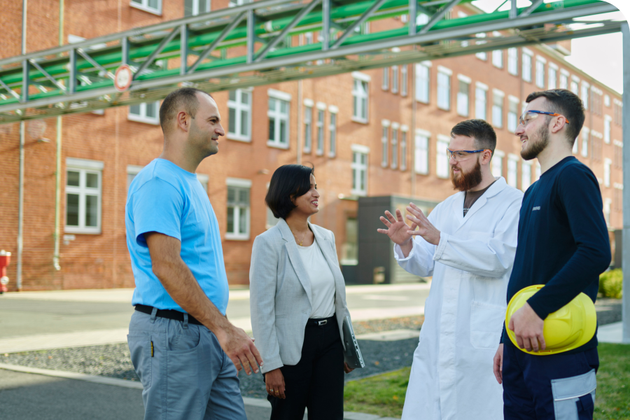 Four professionals, including a woman and three men, converse outdoors near a brick building. One wears a lab coat, another holds a yellow hard hat.