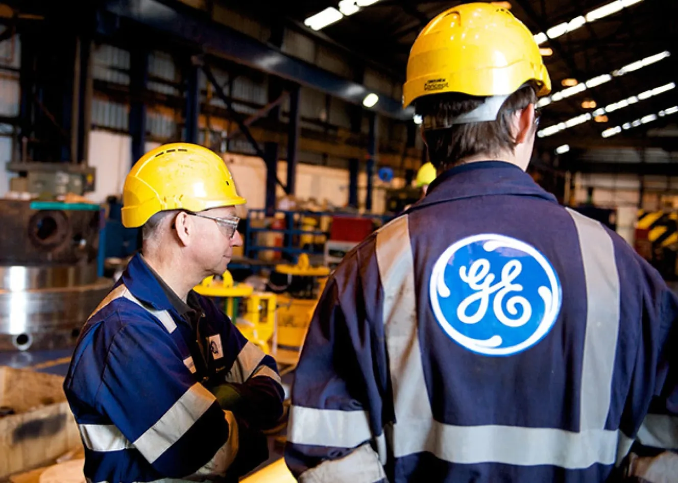 Two workers in blue uniforms and yellow helmets stand in an industrial facility. One has the GE logo on his back. Machinery is visible in the background.