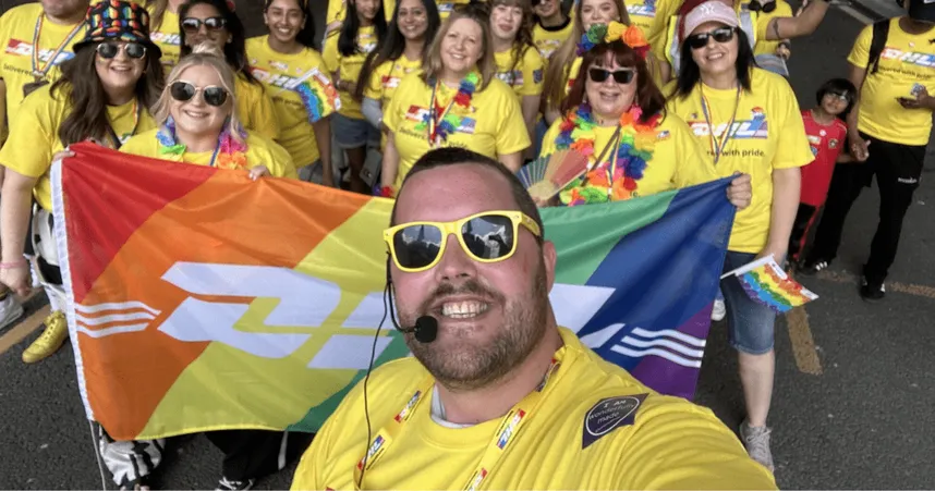 A group of DHL employees dressed in yellow “Deliver with pride” t-shirts gather for a group selfie during a Pride event. The person in front, wearing yellow sunglasses and a headset, smiles while holding a DHL-branded rainbow Pride flag. The group behind is also smiling and waving small Pride flags, celebrating inclusivity and diversity.