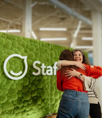 Two Staffbase colleagues sharing a warm hug in front of the green moss logo wall in the office.