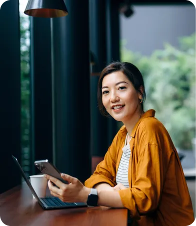 A woman in a casual setting sits at a table with a laptop, holding a phone and smiling at the camera.