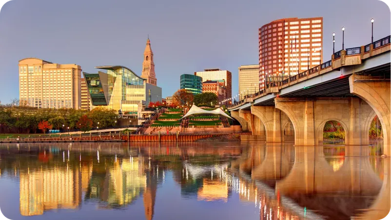 Hartford, Connecticut skyline at dusk, reflected in the river with a bridge in the foreground.