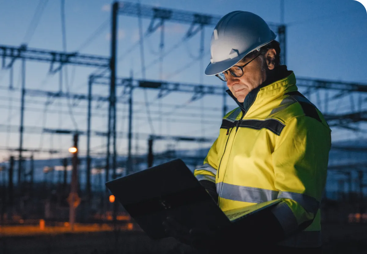 Worker in a high-visibility jacket and hard hat uses a laptop at an industrial site with power lines at dusk.