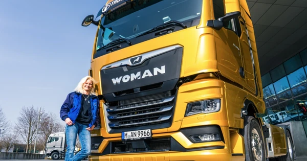 A person in a blue jacket stands beside a large yellow truck with "WOMAN" on the front, parked outdoors near a modern building.