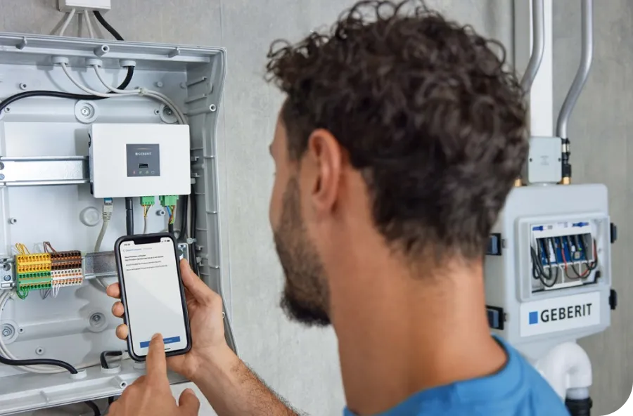 A person uses a smartphone to configure a device inside an open electrical panel labeled "Geberit" on a wall.