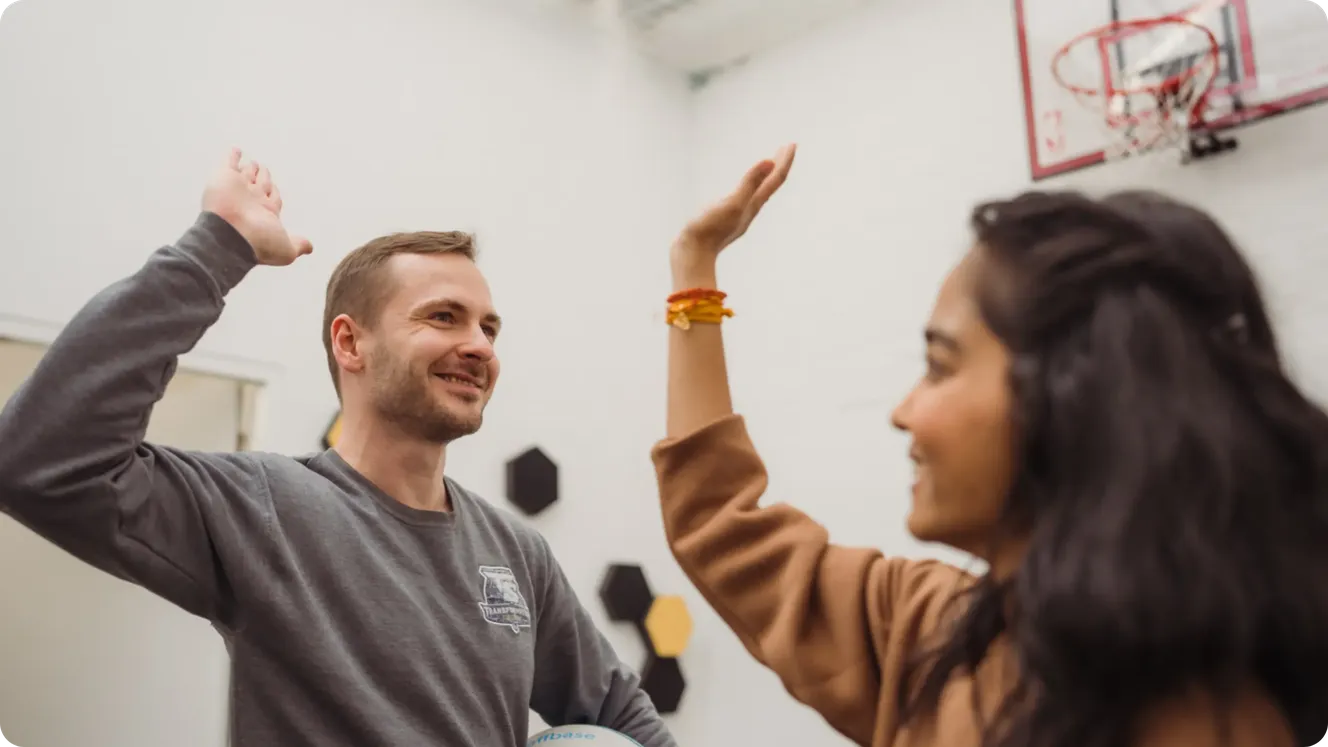 Two Staffbase colleagues high-fiving and smiling in a room with a basketball hoop. Title: Staffbase Colleagues High-Fiving