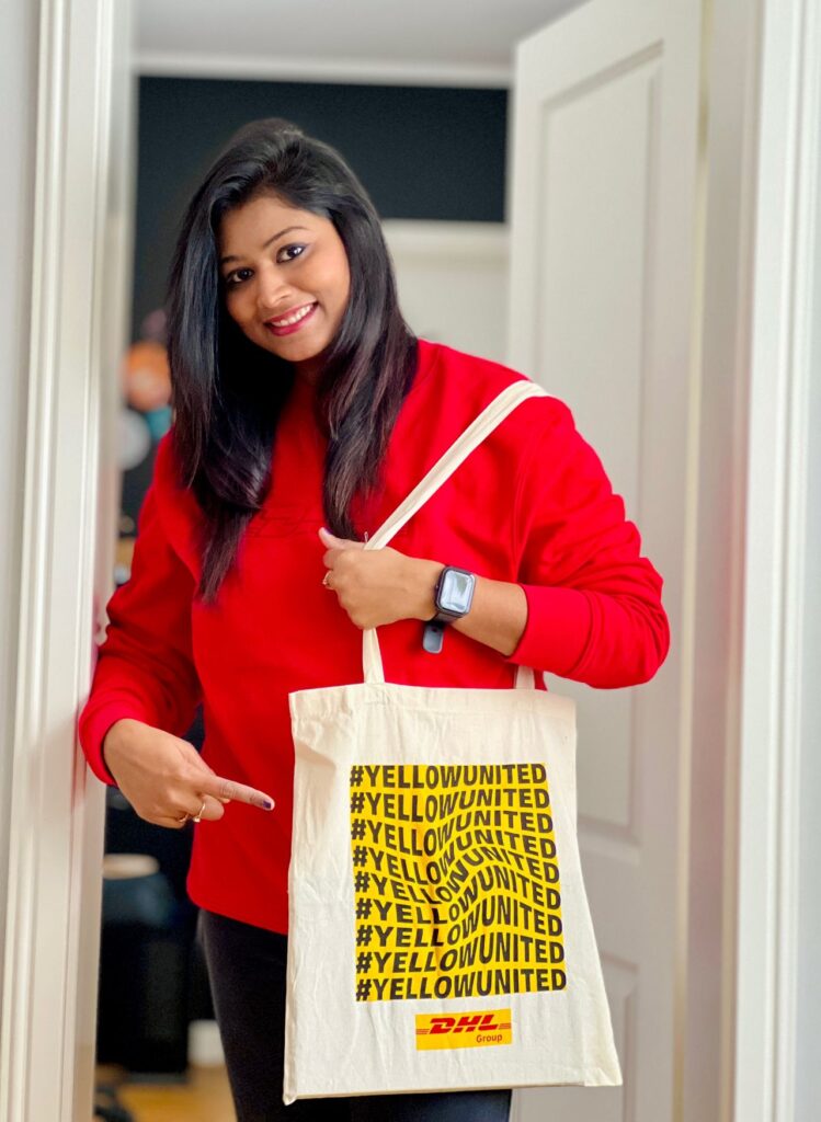 A smiling woman wearing a red sweatshirt stands in a doorway, pointing proudly at a cream-colored tote bag hanging on her shoulder. The tote bag features multiple bold yellow and black “#YELLOWUNITED” hashtags and the DHL Group logo at the bottom. The woman is also wearing a smartwatch.
