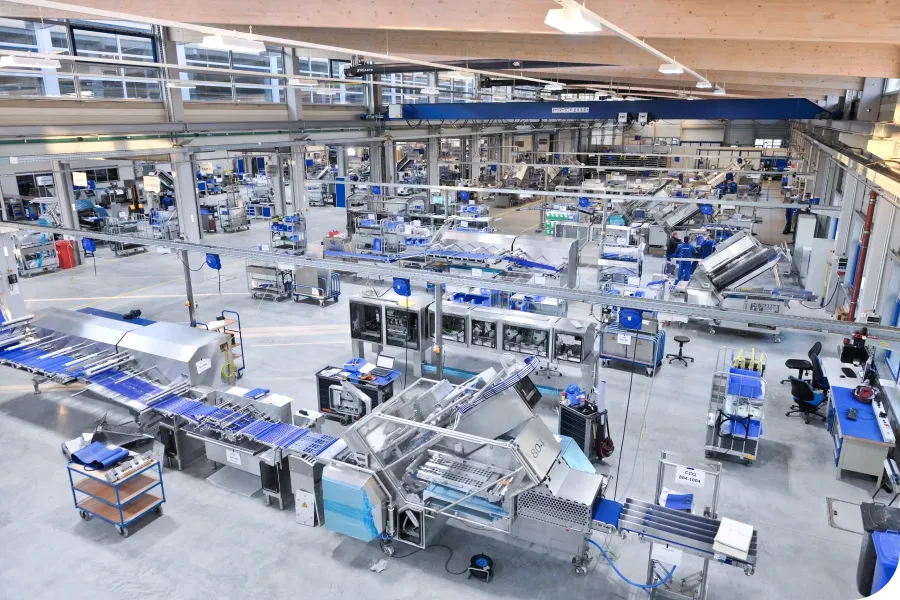 A large, modern factory interior with automated machinery and conveyor belts, featuring blue accents and organized workstations.