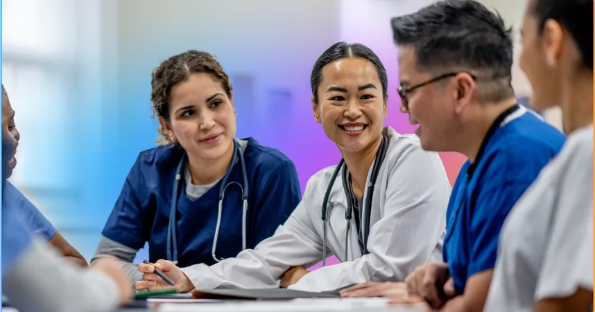 A diverse group of smiling healthcare professionals in scrubs and lab coats having a discussion around a table.