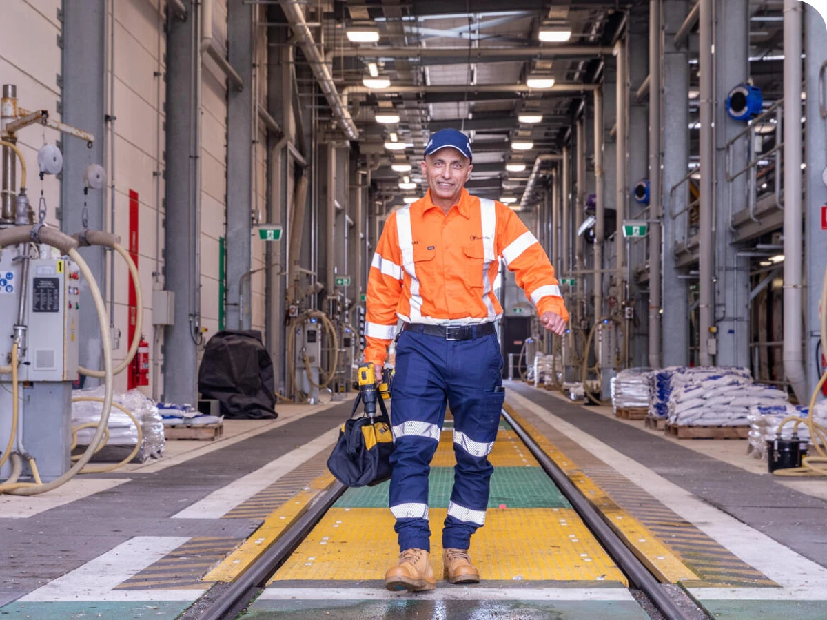 Worker in an orange safety jacket and blue pants walks confidently in an industrial facility, holding a tool bag.