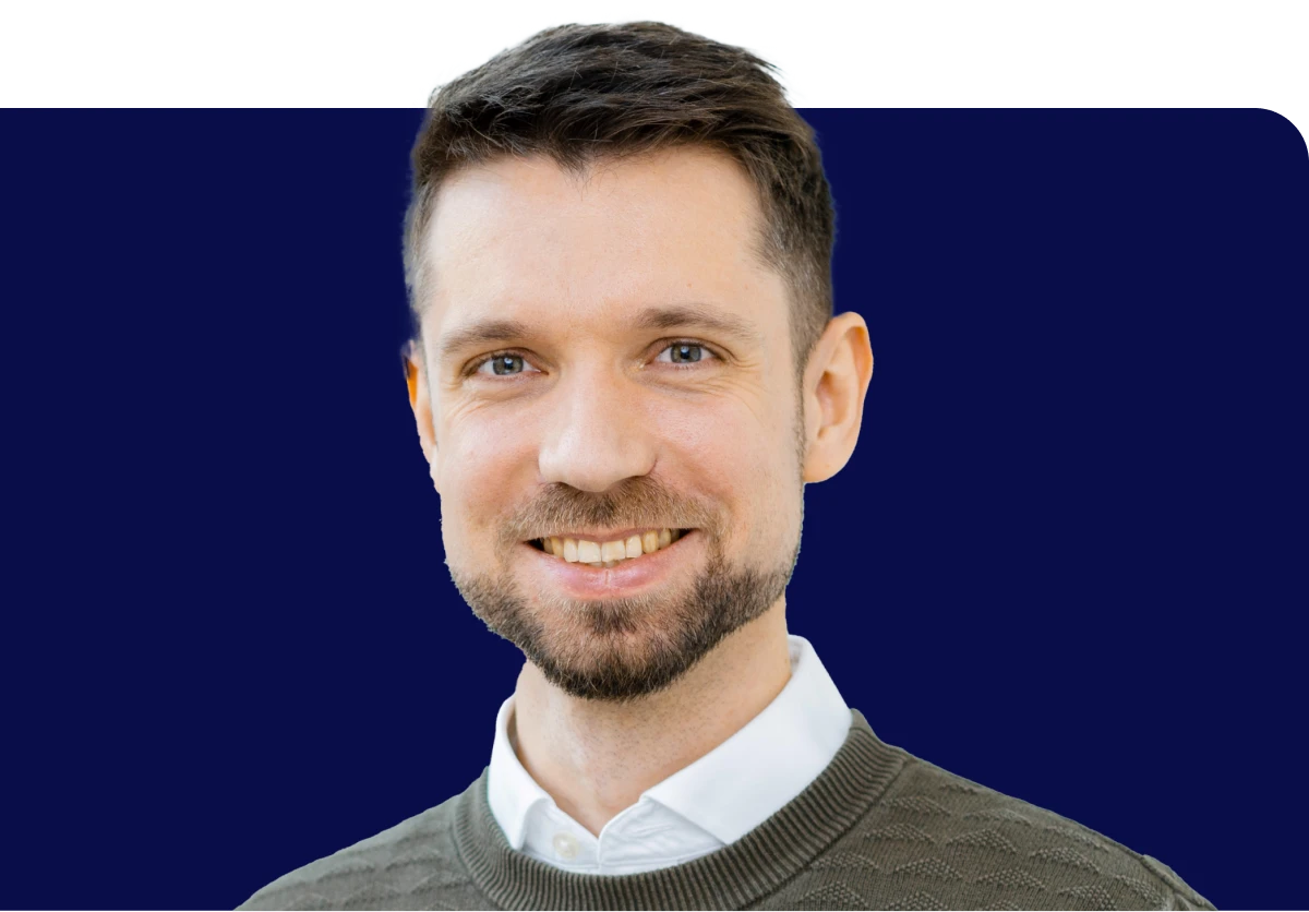 Head of Content & Editorial, Heraeus. Smiling man with short dark hair and beard, wearing a white shirt and gray sweater, against a dark blue background.