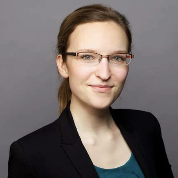 Smiling person with glasses and brown hair, wearing a dark blazer and teal top, against a plain gray background.