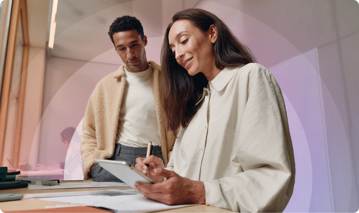 Two people in a modern office setting, one writing on a tablet, the other observing. Both are casually dressed and engaged in discussion.