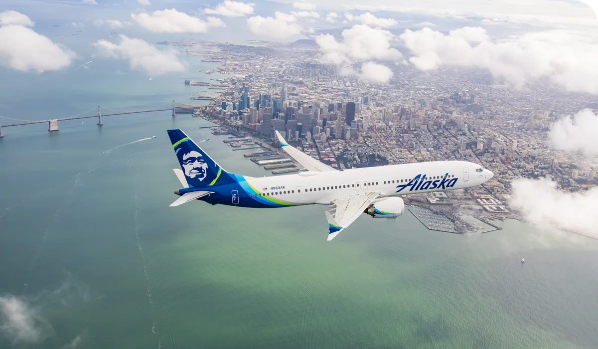 An Alaska Airlines plane flying over a cityscape with a bridge and waterfront below, under a partly cloudy sky.
