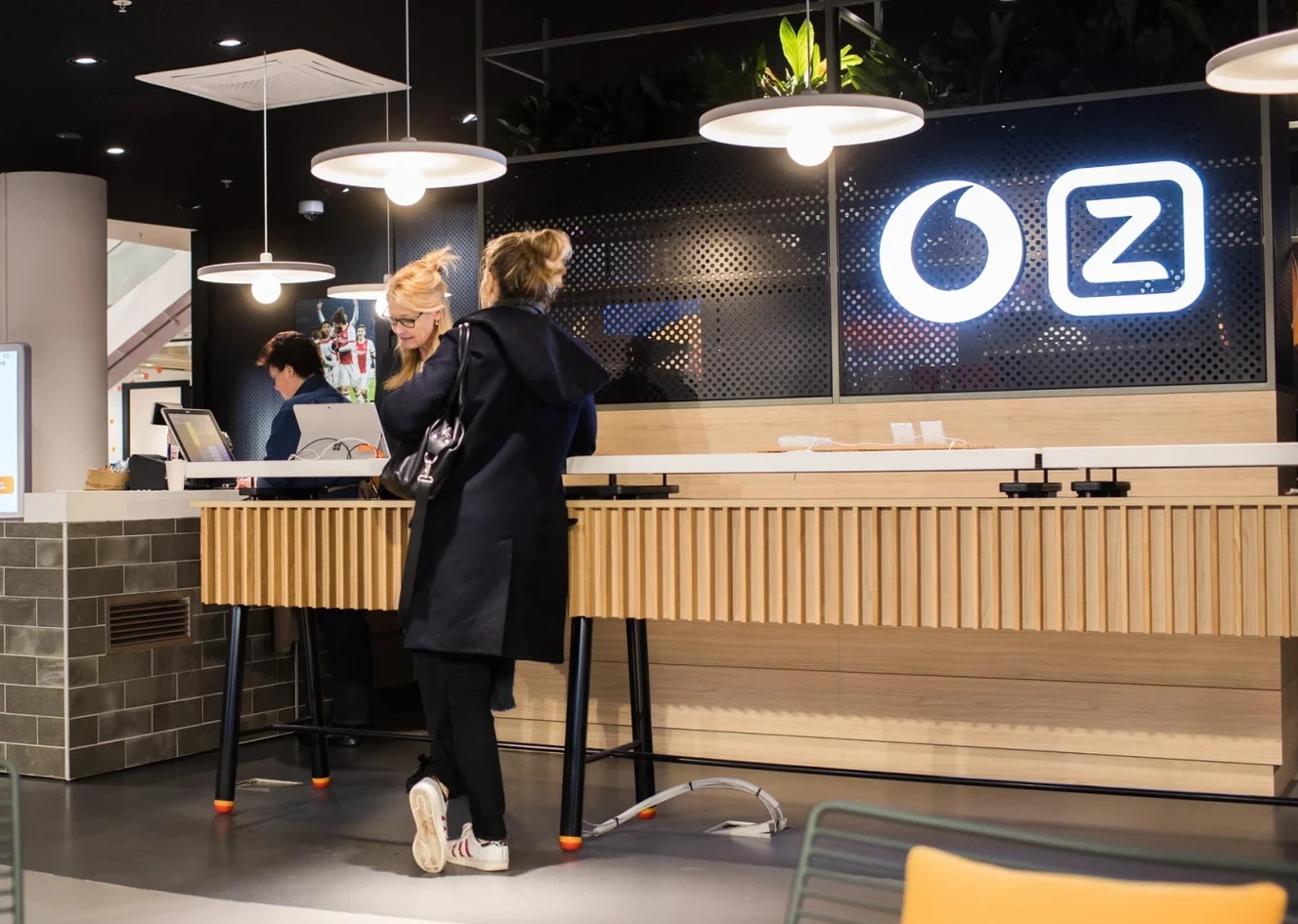 Two people stand at a modern, wooden counter in a well-lit café with hanging lights and a logo on the wall.