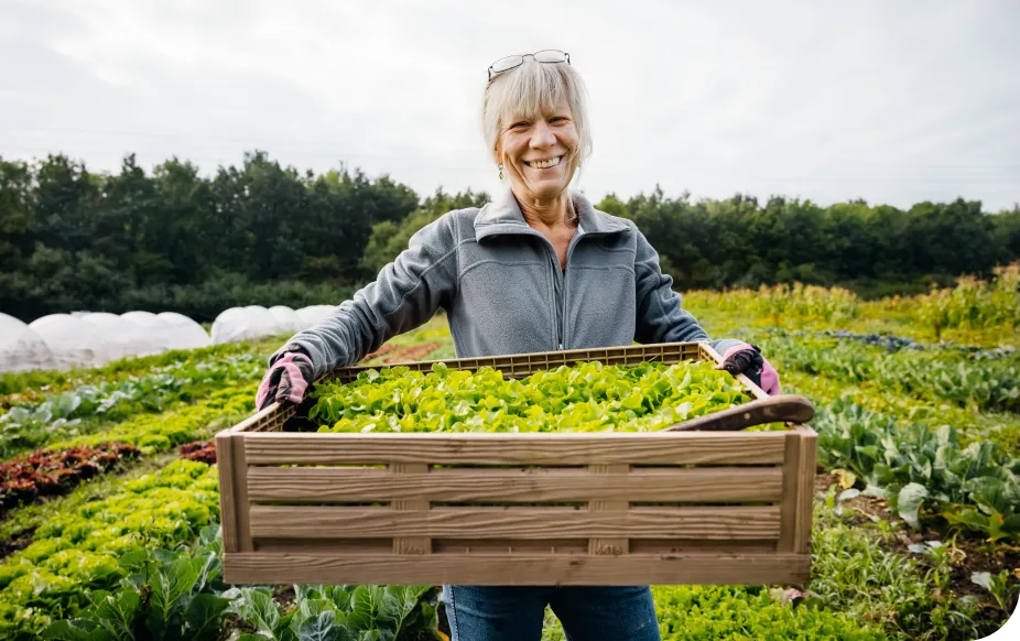 Smiling woman holding a wooden crate full of fresh greens in a lush vegetable garden, wearing gloves and a jacket.
