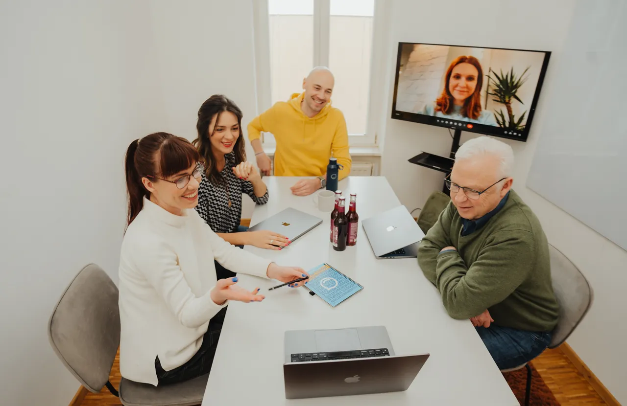 Four people in a meeting room with laptops and drinks, video conferencing with a person on a screen, engaged in discussion.