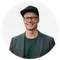 Headshot of a men with a cap, glasses and a grey jacket. he is smiling into the camera.