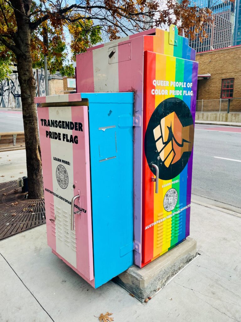 Utility box on a city sidewalk painted with pride flag murals including the Transgender Pride Flag and Queer People of Color Pride Flag with QR codes.