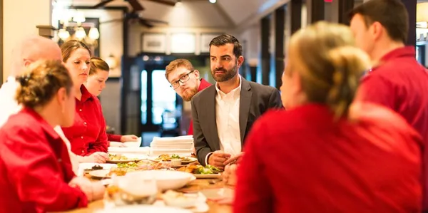 A group of restaurant staff in red shirts listens attentively to a man in a suit during a meeting at a wooden table with food dishes.